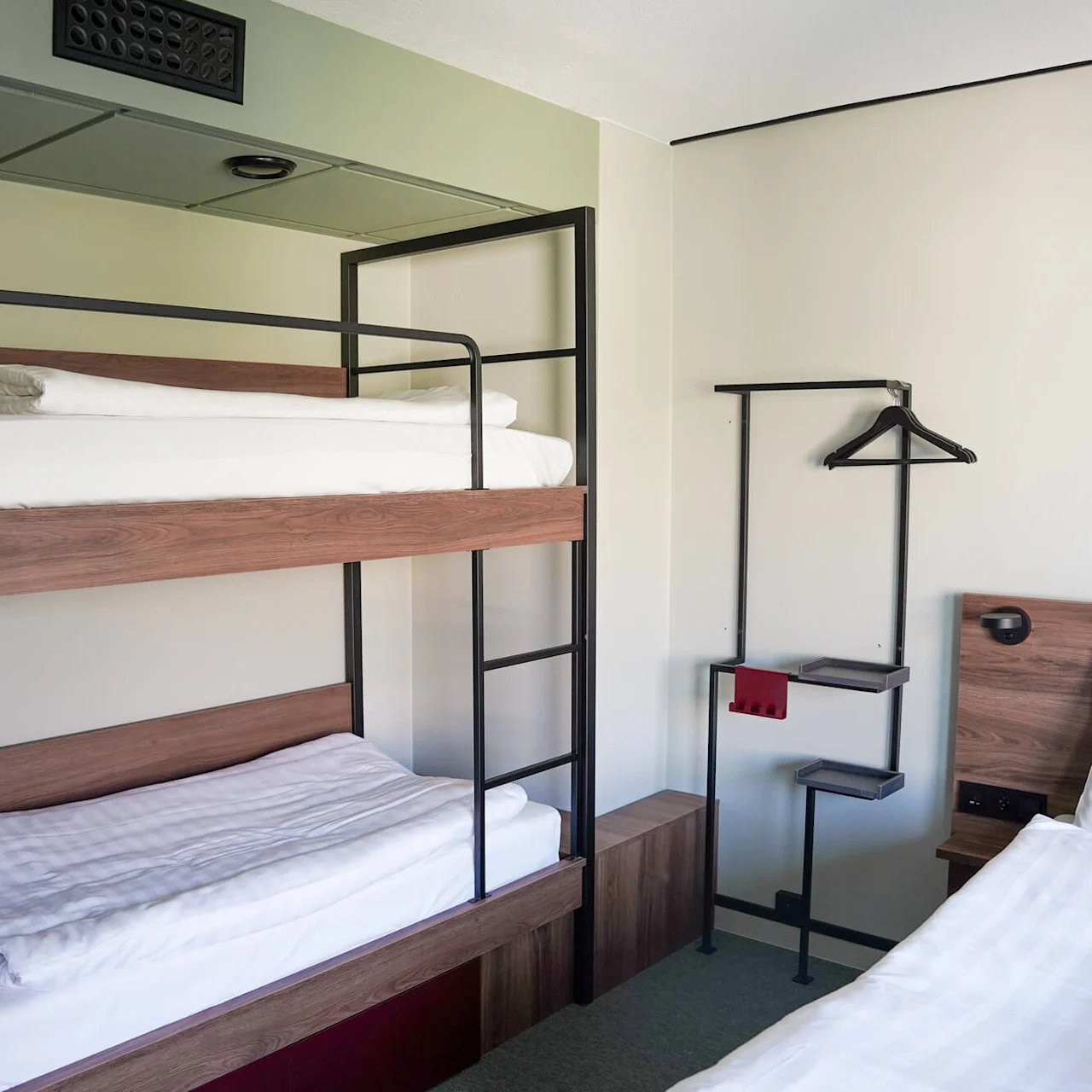 Bunk beds with white bedding are positioned against a pale green wall. A simple storage unit holds hangers and shelves beside the beds.