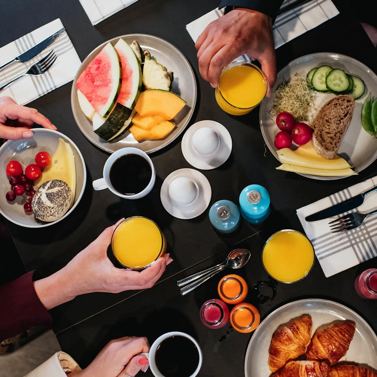 A table displays a breakfast setting with hands reaching for coffee, orange juice, and plates of fruit, bread, cheese, croissants, and yogurt in a casual dining environment.