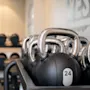 Kettlebells med metalhåndtag står på en rack i træningscenteret. Tallene "24" og "12" er synlige. Håndvægte er lineet op i baggrunden mod en sløret væg.