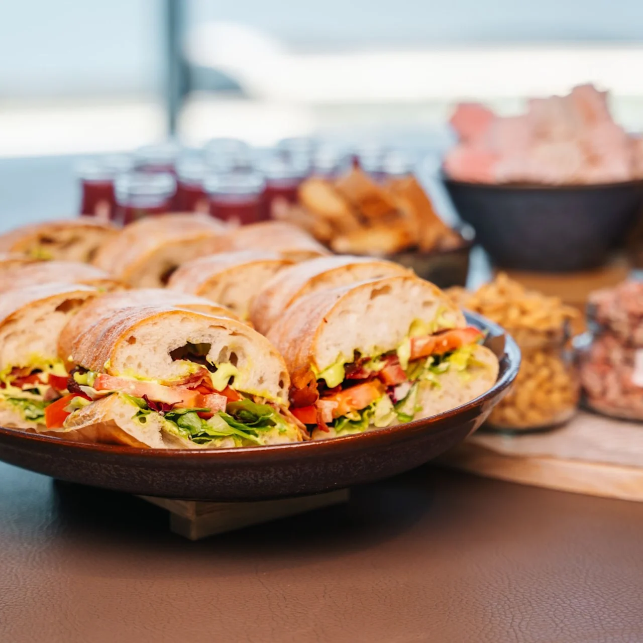Sandwiches with vegetables and meats are arranged on a plate, surrounded by bowls of snacks like nuts and crackers on a table, suggesting a buffet or casual dining setting.