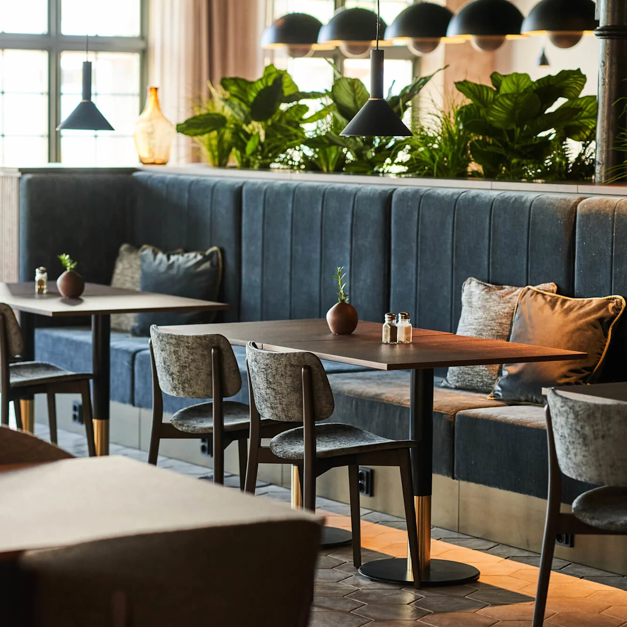 Moderne restaurantborde med grå stole og mørke bænke under hængende lamper; grønne planter dekorerer skillevæggen, og naturligt lys strømmer ind gennem store vinduer.