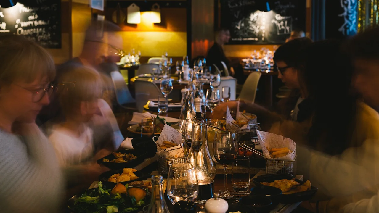 Folk nyder et måltid på en livlig restaurant, med mad og drikke på bordet. En tavle læser After Work.