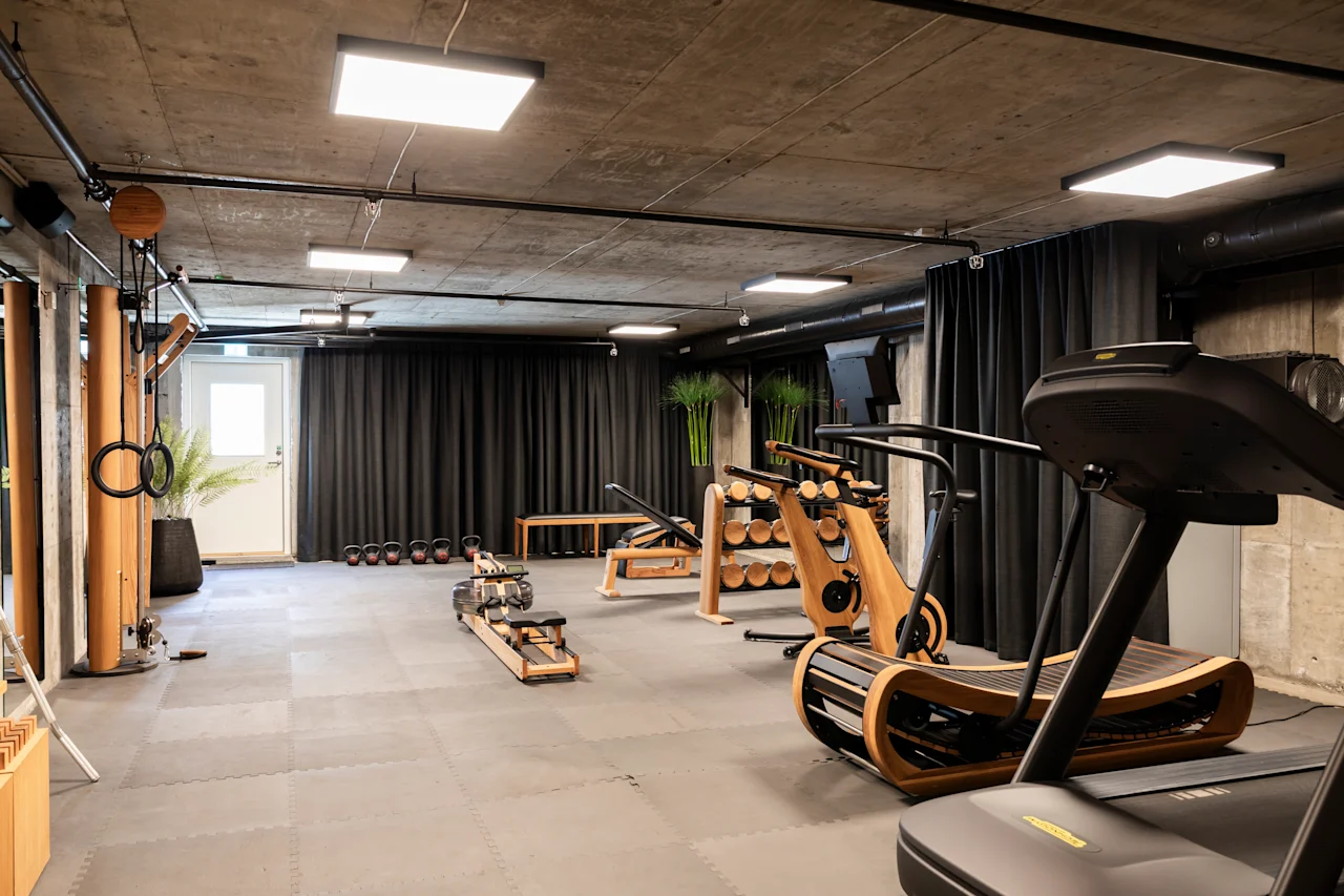 Löpbands står stilla i ett modernt gym med träningsutrustning av trä, mörka gardiner och krukväxter, vilket skapar ett elegant, minimalistiskt träningsutrymme.