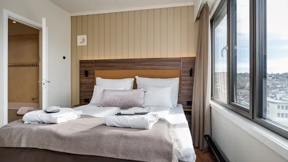 A neatly made bed with white sheets and pillows is adorned with folded towels. It rests in a sunlit room with large windows overlooking a cityscape.
