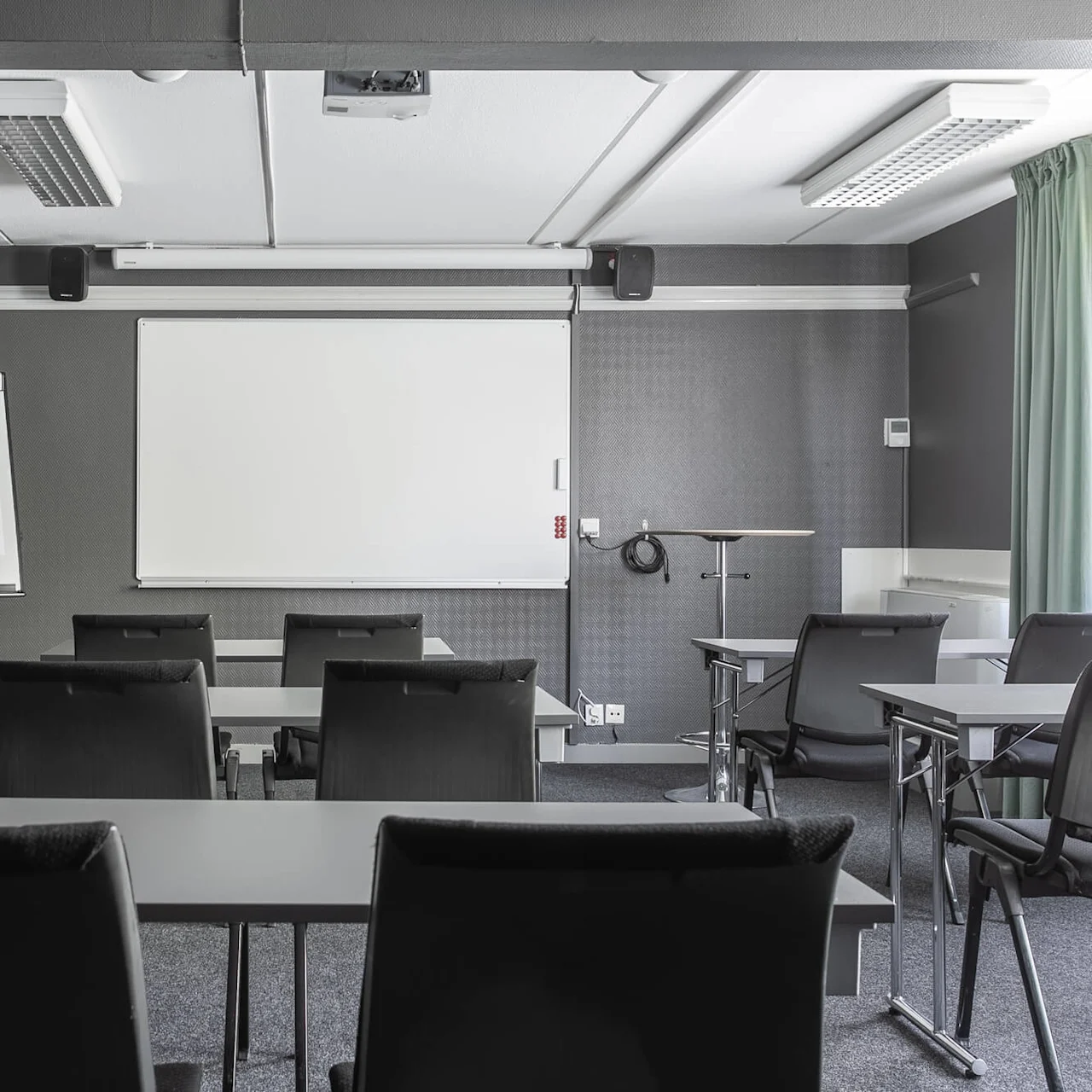 Tomt konferencelokale med sorte stole og borde vendt mod en whiteboard. En flipover står til venstre, og der er en vægur over. Rummet har grå vægge og tæppe.