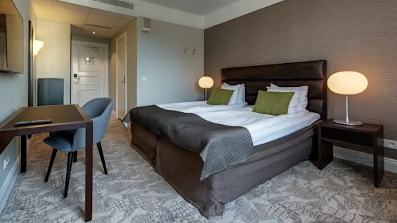 Et lyst hotellrom med en stor seng, skrivebord og stol, og dempet innredning i jordfarger.