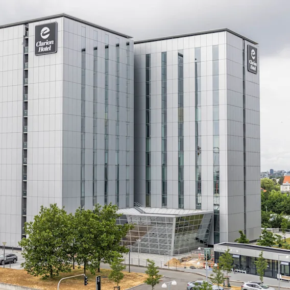 En høj, moderne bygning mærket “Clarion Hotel” med store glasvinduer står i et urbant miljø. Flagene vejrer nær indgangen.