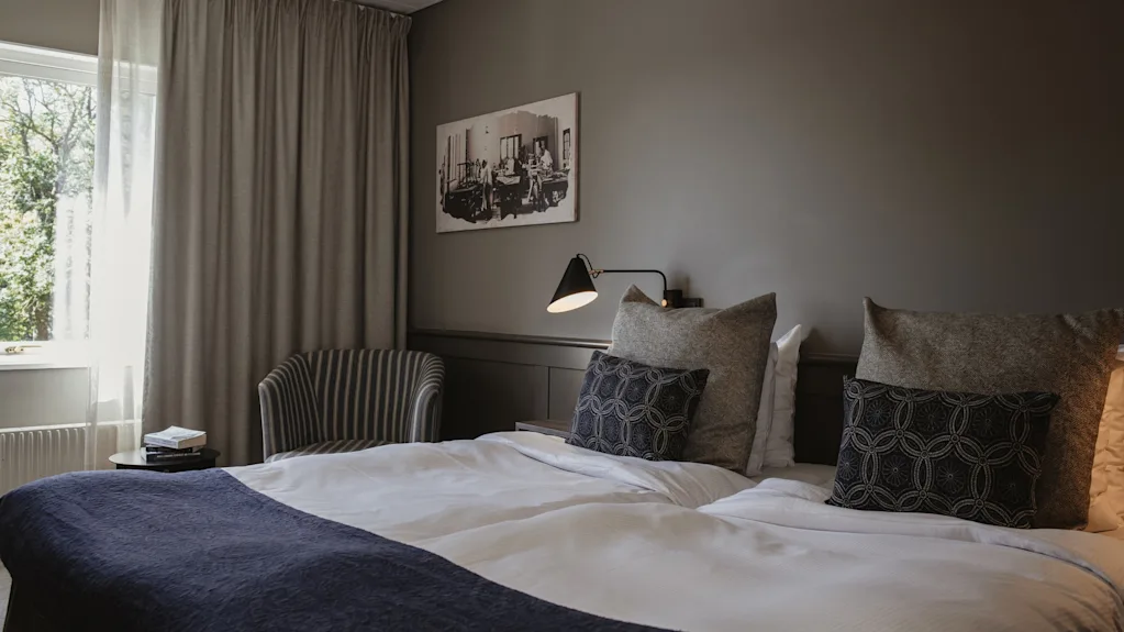 Ett mysigt hotellrum med en prydligt bäddad säng, en bekväm stol vid fönstret och en varm sänglampa.