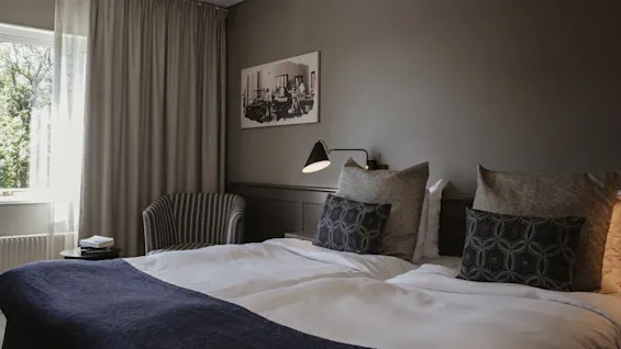 Ett mysigt hotellrum med en prydligt bäddad säng, en bekväm stol vid fönstret och en varm sänglampa.