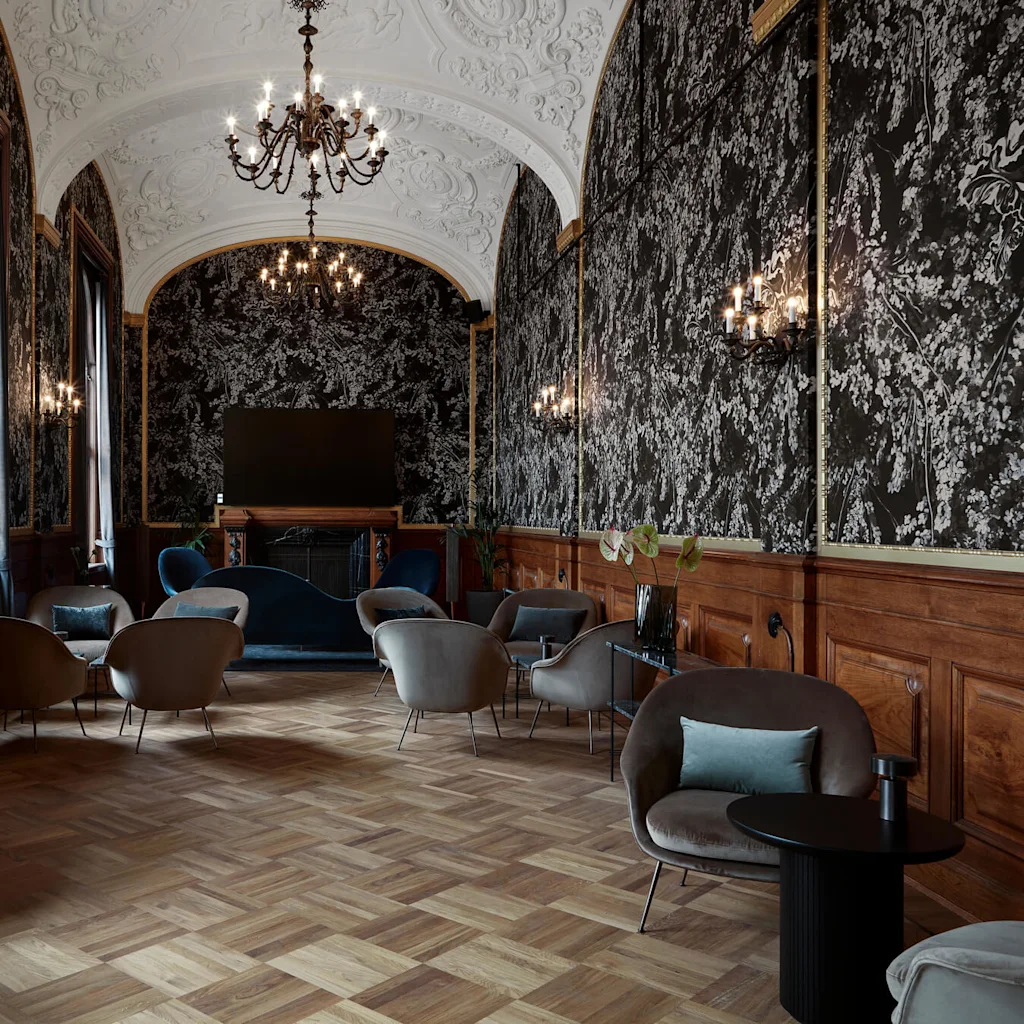 Elegante loungestole omgiver mørke sofaborde under udsmykkede lysekroner i et overdådigt rum med indviklede tapeter og parketgulv.