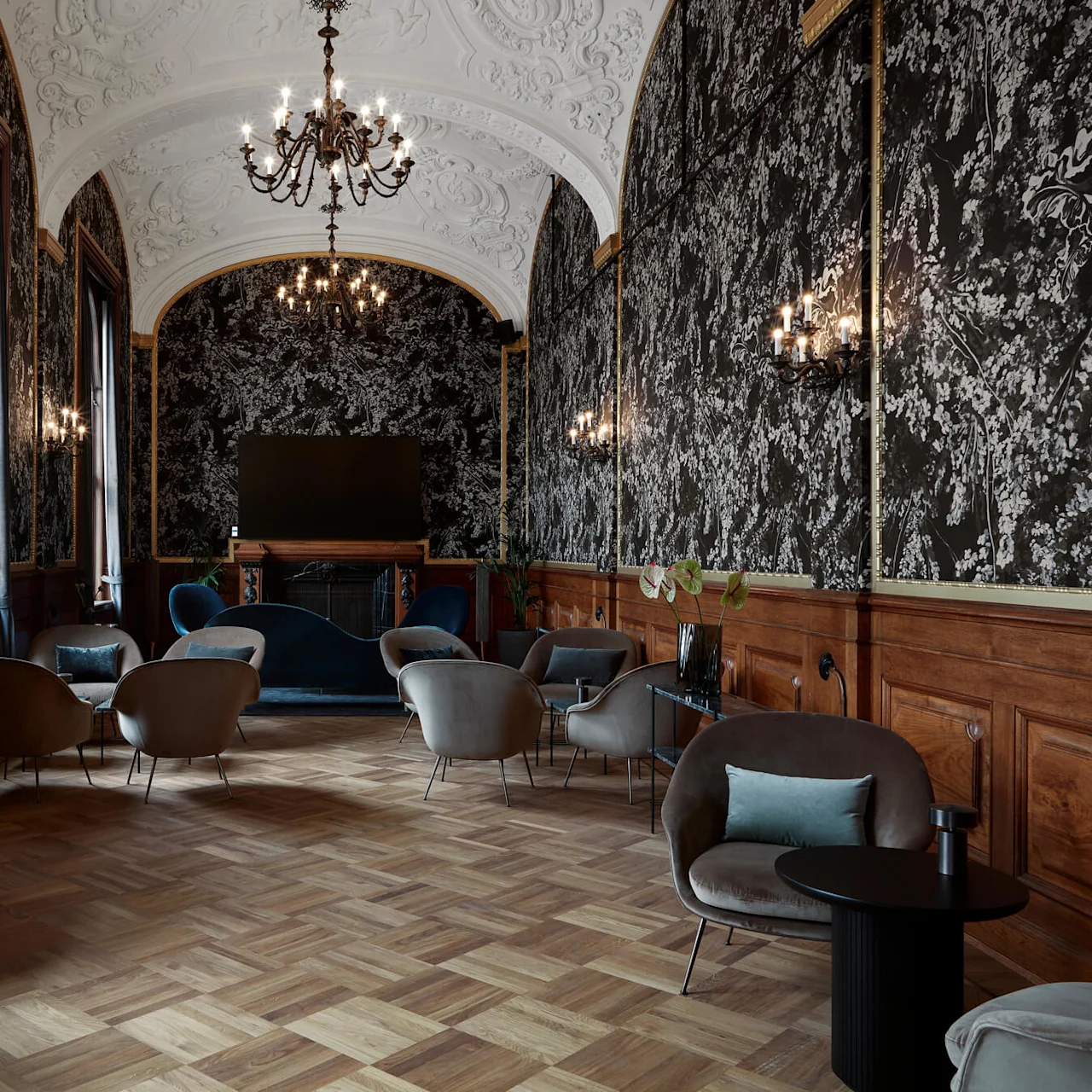 Elegant lounge chairs encircle dark coffee tables underneath ornate chandeliers in a lavish room with intricate wallpaper and parquet flooring.