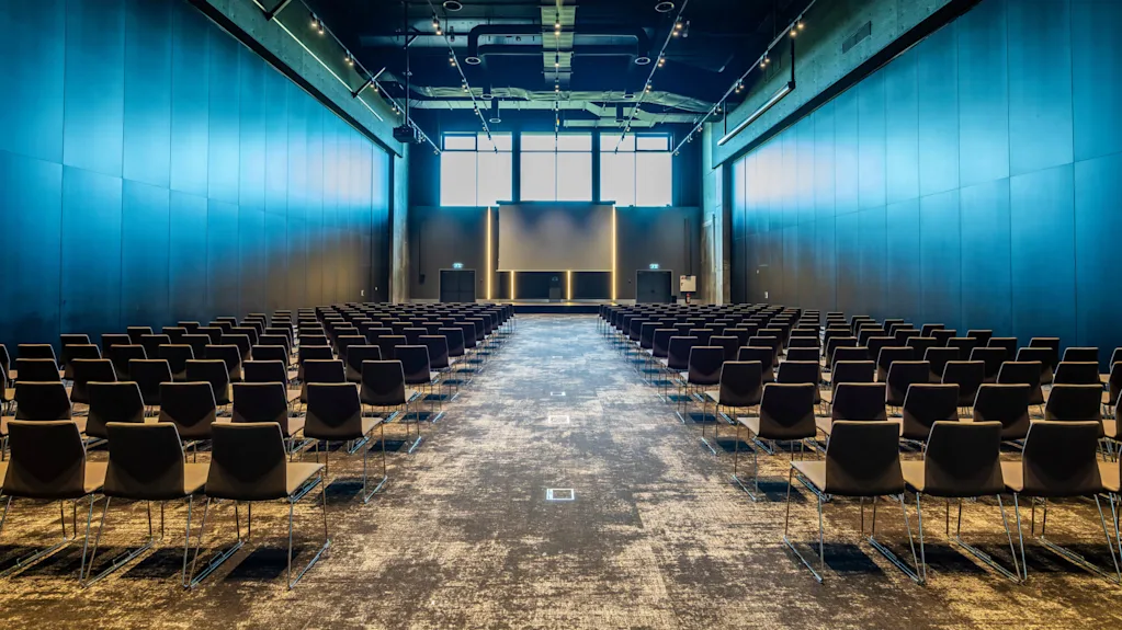 Rows of empty black chairs face a large screen in a spacious conference room illuminated by overhead lights. High windows allow natural light to enter.