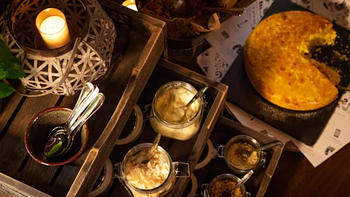 A rustic meal setup, candles providing ambiance, with a pie missing a slice on a wooden table filled with condiments.