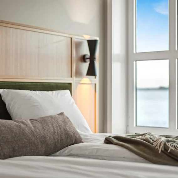 Et koselig hotellrom med en seng, puter og et grønt teppe, ved siden av et vindu med utsikt over sjøen og himmelen.