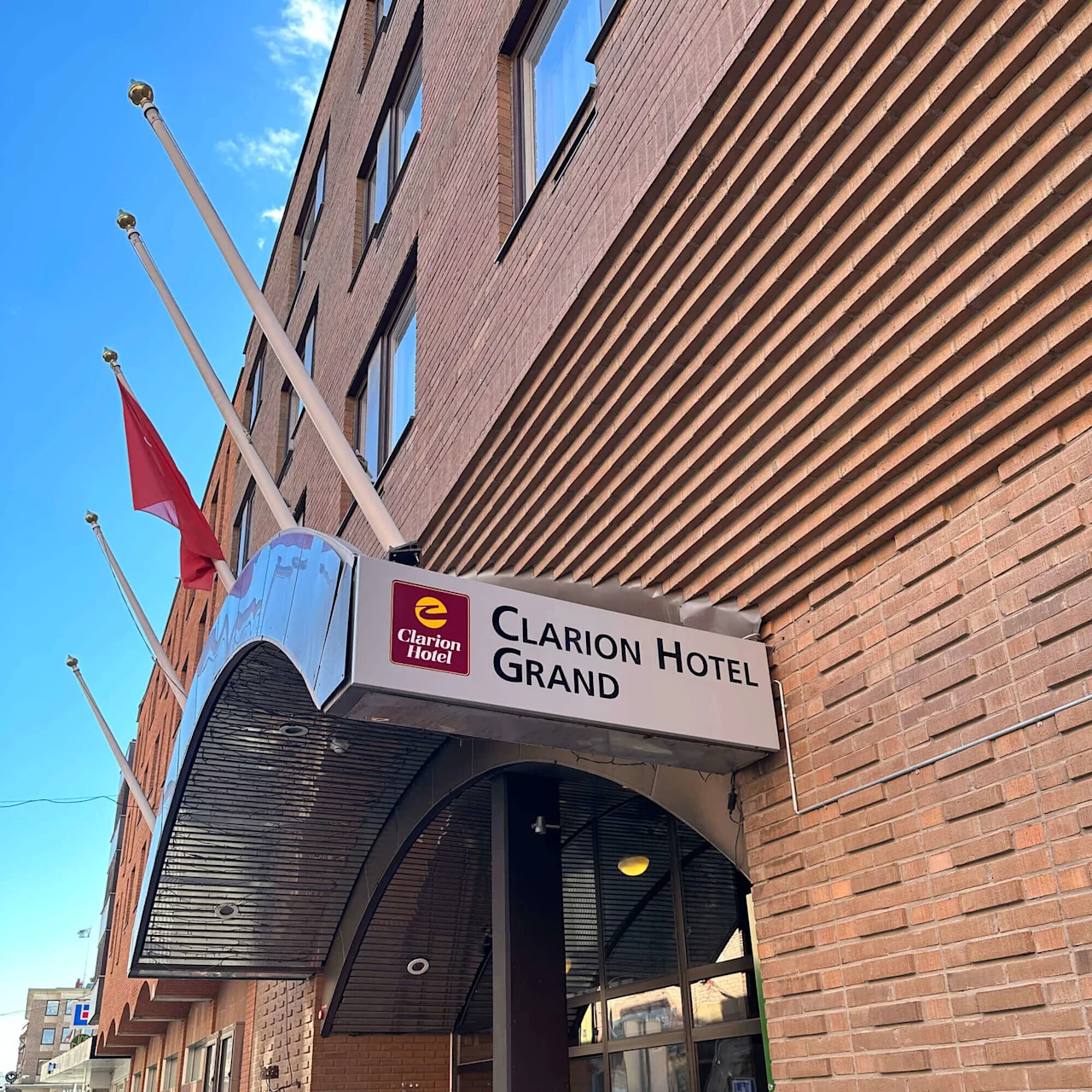 En hotellentré med en skylt som lyder "Clarion Hotel Grand." Flera flaggstänger reser sig från byggnadens tegelfasad i en stadsmiljö.