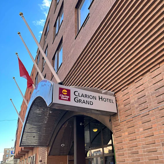 En hotellentré med en skylt som lyder "Clarion Hotel Grand." Flera flaggstänger reser sig från byggnadens tegelfasad i en stadsmiljö.