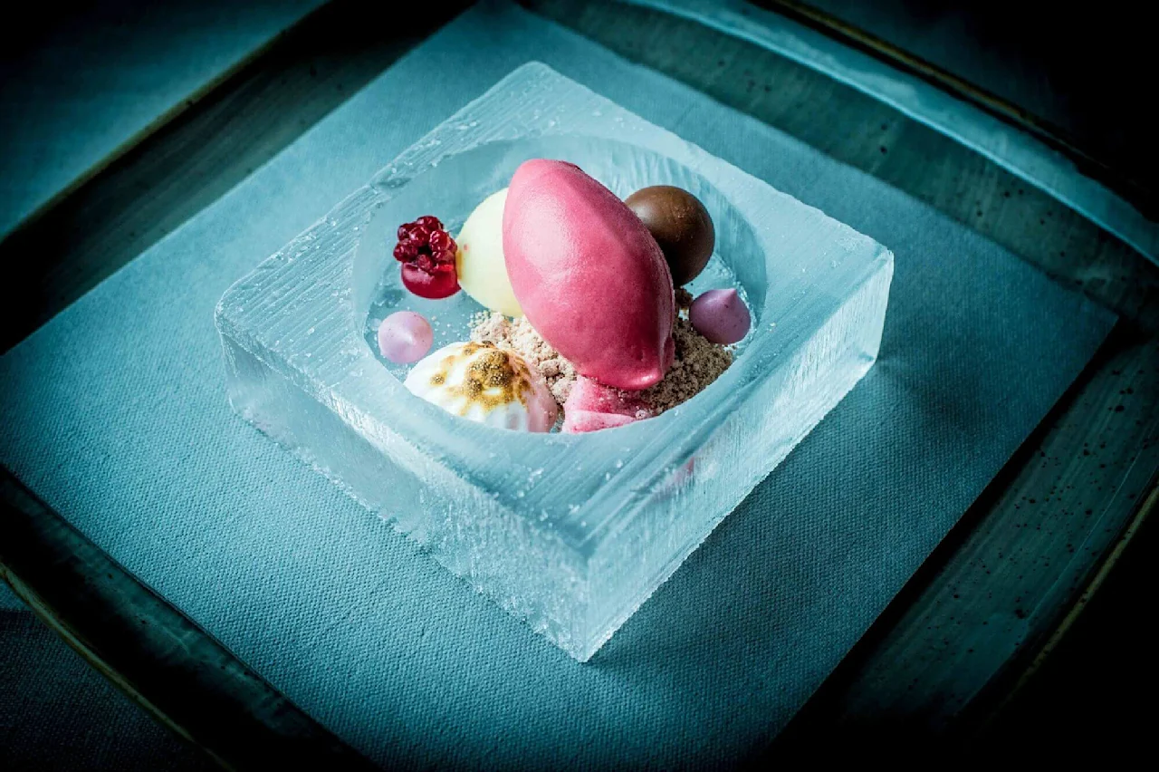 En farverig dessert med sorbet, mousse og bær arrangeret i en udhugget isbowle, placeret på en blå stofovertrukket overflade.