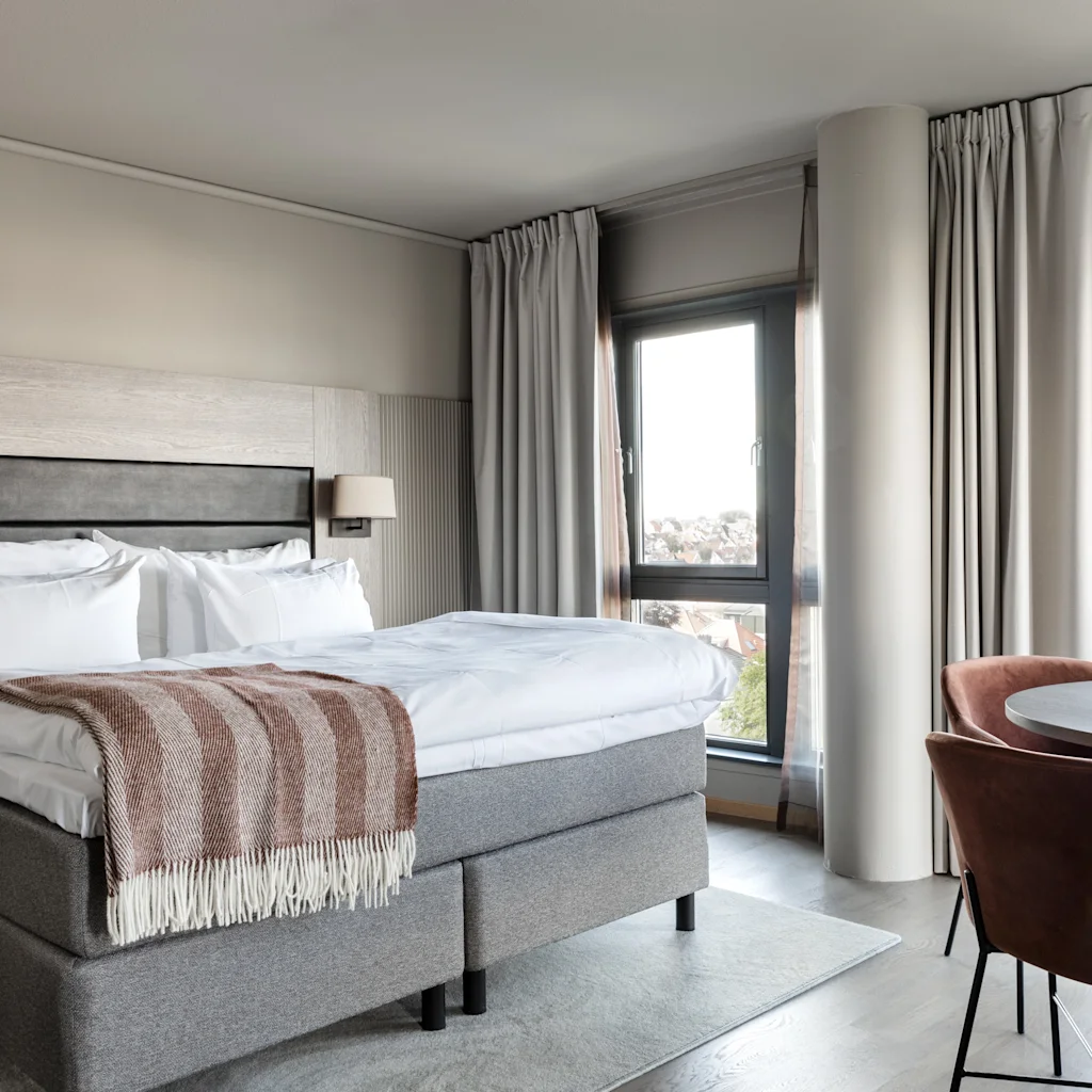 A bright and inviting hotel room featuring a large bed with a cozy throw, a small dining area, and expansive windows.