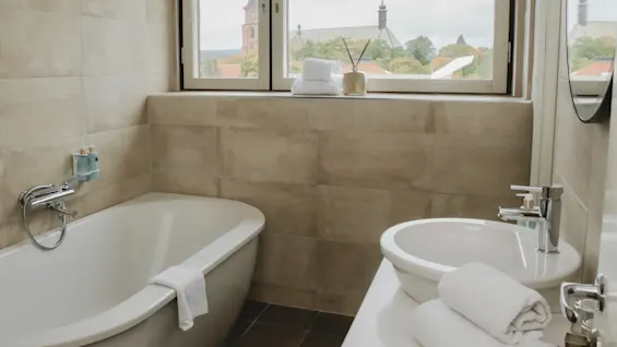 Bathtub with a towel on it and a sink beside, in a tiled bathroom with a window showing church spires and rooftops outside.