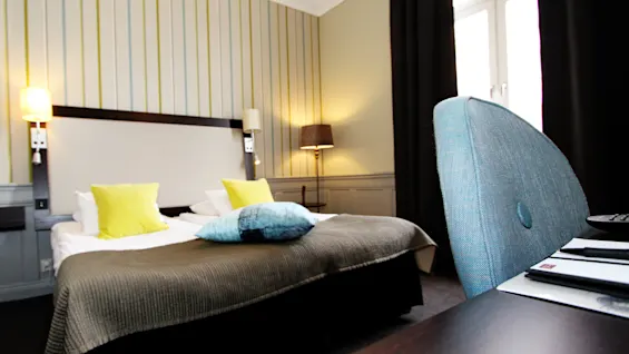 A cozy hotel room features a neatly made double bed with yellow and blue pillows, lamps on either side. Striped wallpaper and dark curtains enhance the space's modern look.