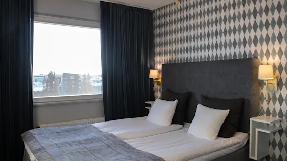 A neatly made bed with dark headboard and white pillows stands against a diamond-patterned wallpaper. Two wall lamps illuminate the scene. A window with dark curtains overlooks a snowy urban setting.