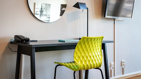A lime green geometric chair is pushed under a sleek black desk equipped with a telephone, a blue notebook, and a modern lamp. A round mirror and wall-mounted TV are nearby.