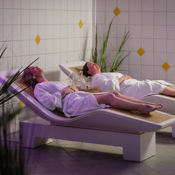 To kvinner i hvite badekåper som slapper av på ergonomiske liggestoler i et fredelig spa-område.
