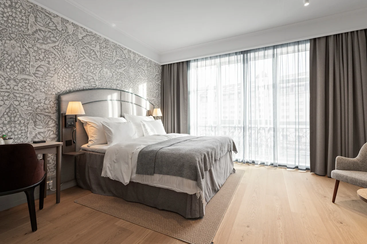 A bright and inviting hotel room with a large bed, elegant patterned wallpaper, and sheer curtains letting in natural light.