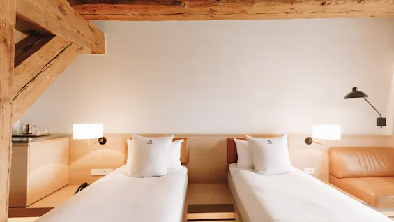Ljust hotellrum med två enkelsängar, synliga träbjälkar i taket och mysig belysning.
