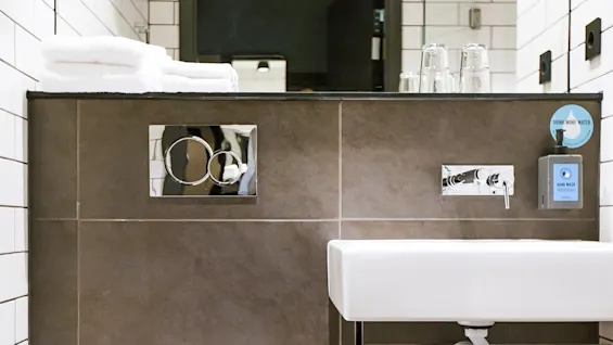 A modern bathroom features a white toilet and sink with dark tiles. Towels, glasses, and soap are on a shelf. A sign reads, "DRINK MORE WATER HAND WASH."