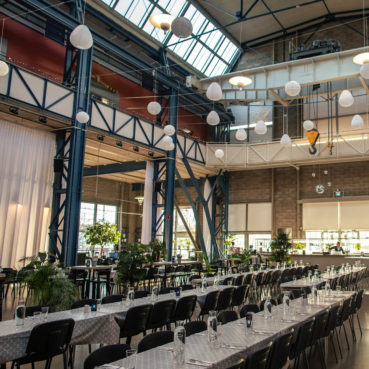 Tables set with glasses and cutlery, arranged in an industrial-style venue with high ceilings and hanging lights. Large windows illuminate the space, surrounded by steel beams and green plants.