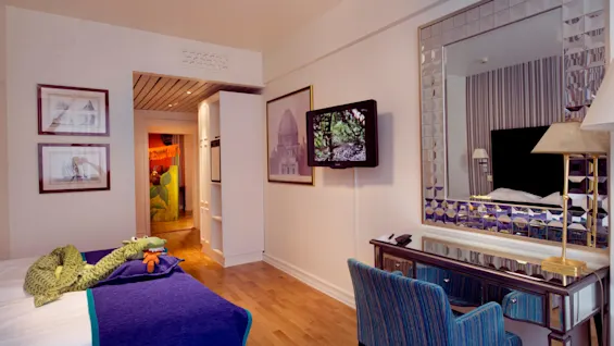Et pent arrangert hotellrom med en myk krokodille leke på sengen, vegghengt TV, dekorativt speil og innrammet kunst.