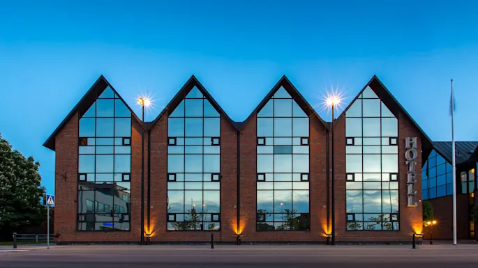 Et moderne hotell med store vinduer og trekantede topper i skumringen, med tent lys og klar blå himmel.