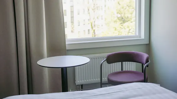 Et lite rundt bord og en lilla stol er plassert ved et stort vindu med utsikt over trær og en bygning, i et mykt opplyst rom med beige gardiner.