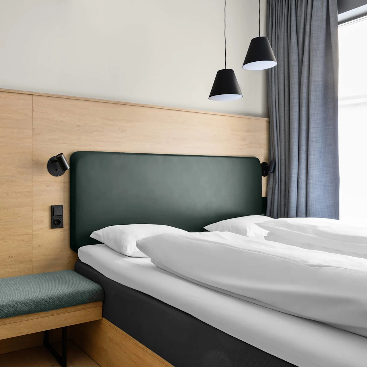 A neatly made bed with white linens against a wood-paneled wall. A green cushioned headboard sits beneath two black pendant lights. Gray curtains cover a window on the right.