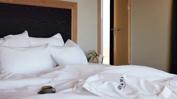 Bed with white pillows and duvet, featuring a wooden headboard. Robe with "WOOD hotel" text on the bed, and a small cup beside it. Door slightly open in a cozy room.