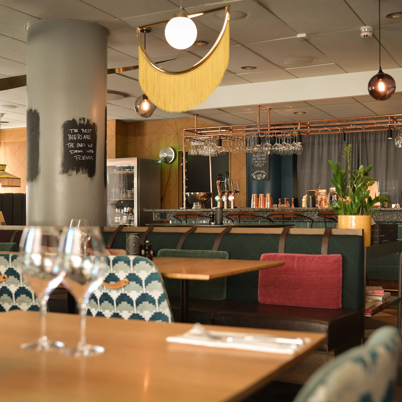 Vinglas står på et træbord i en stilfuld restaurant, med moderne indretning med mønstrede stole, hængende lamper og en synlig bardisk. Tekst på søjlen: "THIS WAY FOR GOOD TIMES."