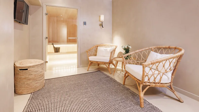 Rattan armchairs and table with a sauna door in the background, in a relaxing, warmly lit spa room.