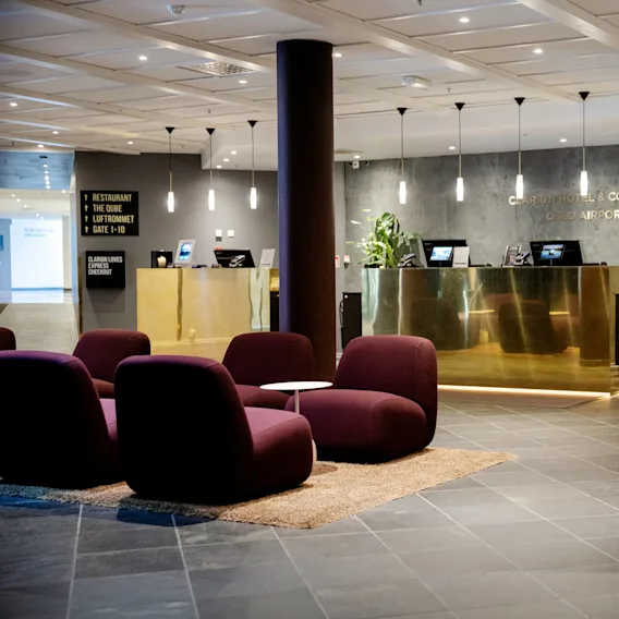 Reception desk in a hotel lobby with gold accents and purple chairs; signs indicate "Restaurant The Qube Luftrommet Gate 1-10" and "Clarion Hotel & Congress Oslo Airport."