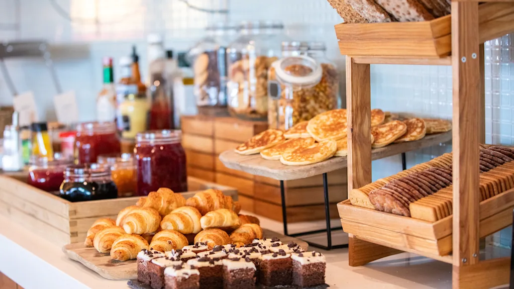 Bakverk och bröd är arrangerade på ett bord, omgivet av sylt- och flingburkar, vilket skapar en frukostbuffé i ett ljust och organiserat köksmiljö.