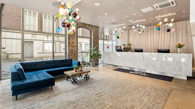 A marble reception desk with colorful pendant lights overhead stands in a modern lobby featuring a blue sectional sofa and a wooden coffee table. "HAVE A GREAT STAY" text on floor mat.