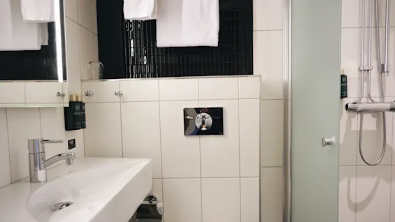 Baderom med hvit vask, kromarmatur og toalett under et hengende håndkle. Svarte fliser står i kontrast til hvite vegger. En dusj med glassdør er til høyre.