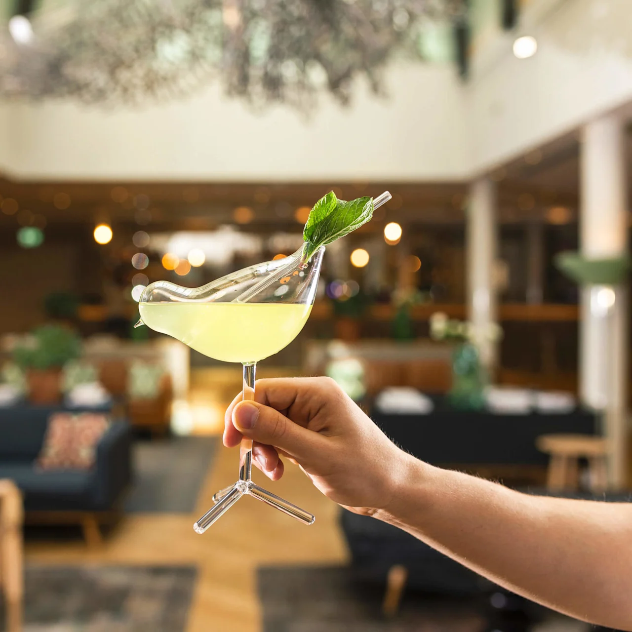 En hånd præsenterer en cocktail med et myntebladsgarniture, sløret baggrund af en stilfuld bar.