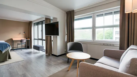A beige sofa and round wooden table are placed in a modern hotel room. Large windows let in natural light, and a wall-mounted TV faces the sofa. A desk is visible in the background.