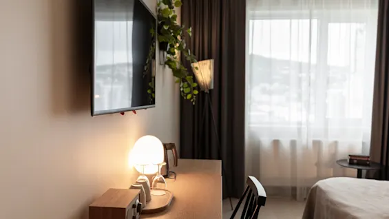 Viihtyisä hotellihuone, jossa on televisio, työpöytä lämpimällä lampulla ja ikkuna, josta avautuu näkymä, luoden rentouttavan tunnelman.