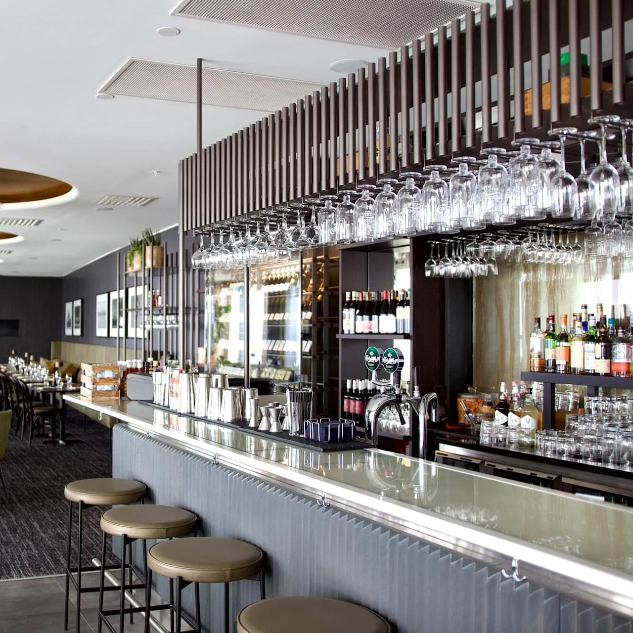 Bar with glass countertops and stools, displaying hanging glasses and various bottles. In the background, elegant dining tables are set in a stylish restaurant with modern decor.