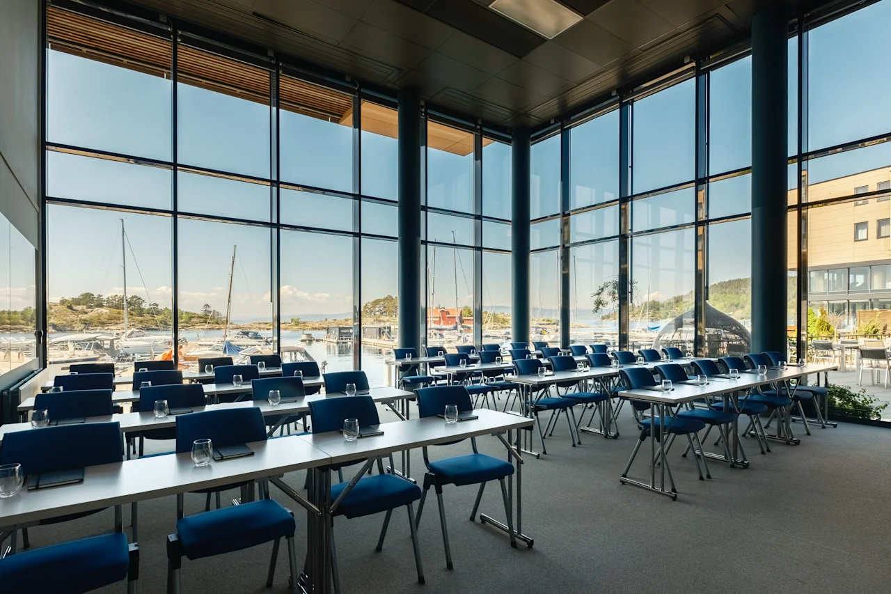 Et lyst konferencelokale med rækker af blå stole og borde, med udsigt over en solrig havn fyldt med både.