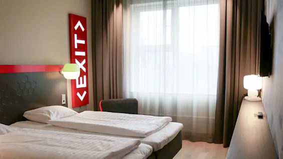 Et moderne hotellrom med to enkeltsenger, et stort opplyst 'EXIT'-skilt over hodegjerdet, og nøytrale gardiner.
