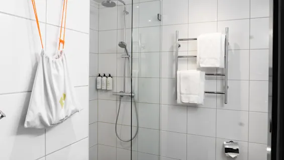 Dusch med kromdetaljer och monterade flaskor är inramad av glas i ett vitt kaklat badrum. Handdukar hänger på en handdukstork. En vit väska med orange snören hänger på krokar.