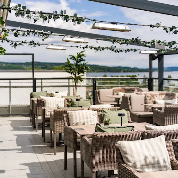 Kurvstoler med puter står på en takterrasse, under en pergola pyntet med lys og grønt. Bakgrunnen viser en naturskjønn utsikt over fjerne trær og vann.
